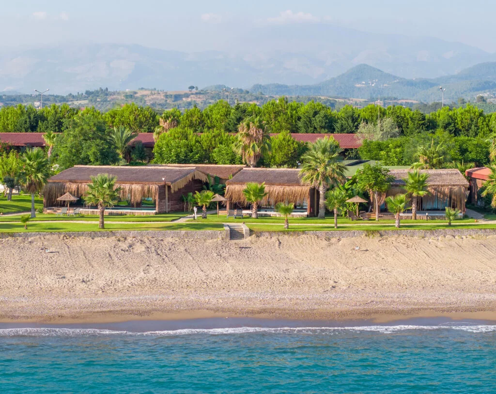 A scenic coastal snapshot of Flora Garden Beach Hotel, with its distinctive thatched-roof structures nestled among lush palm trees, fronting a pebbled beach and the serene blue waters. The backdrop features rolling hills under a soft blue sky, adding to the peaceful and picturesque resort setting.